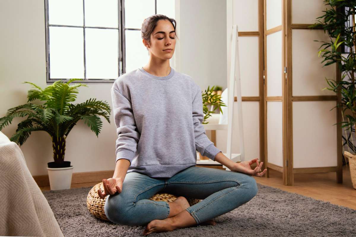 A woman sits cross-legged on a rug in a cozy, well-lit room, meditating with her eyes closed and hands resting on her knees in a relaxed pose